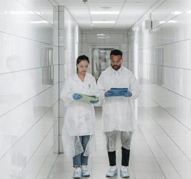 Two scientists in protective gear examine documents in a bright, sterile laboratory hallway.