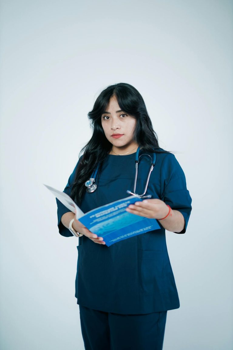 Female nurse in blue scrubs reading a medical book with a stethoscope.