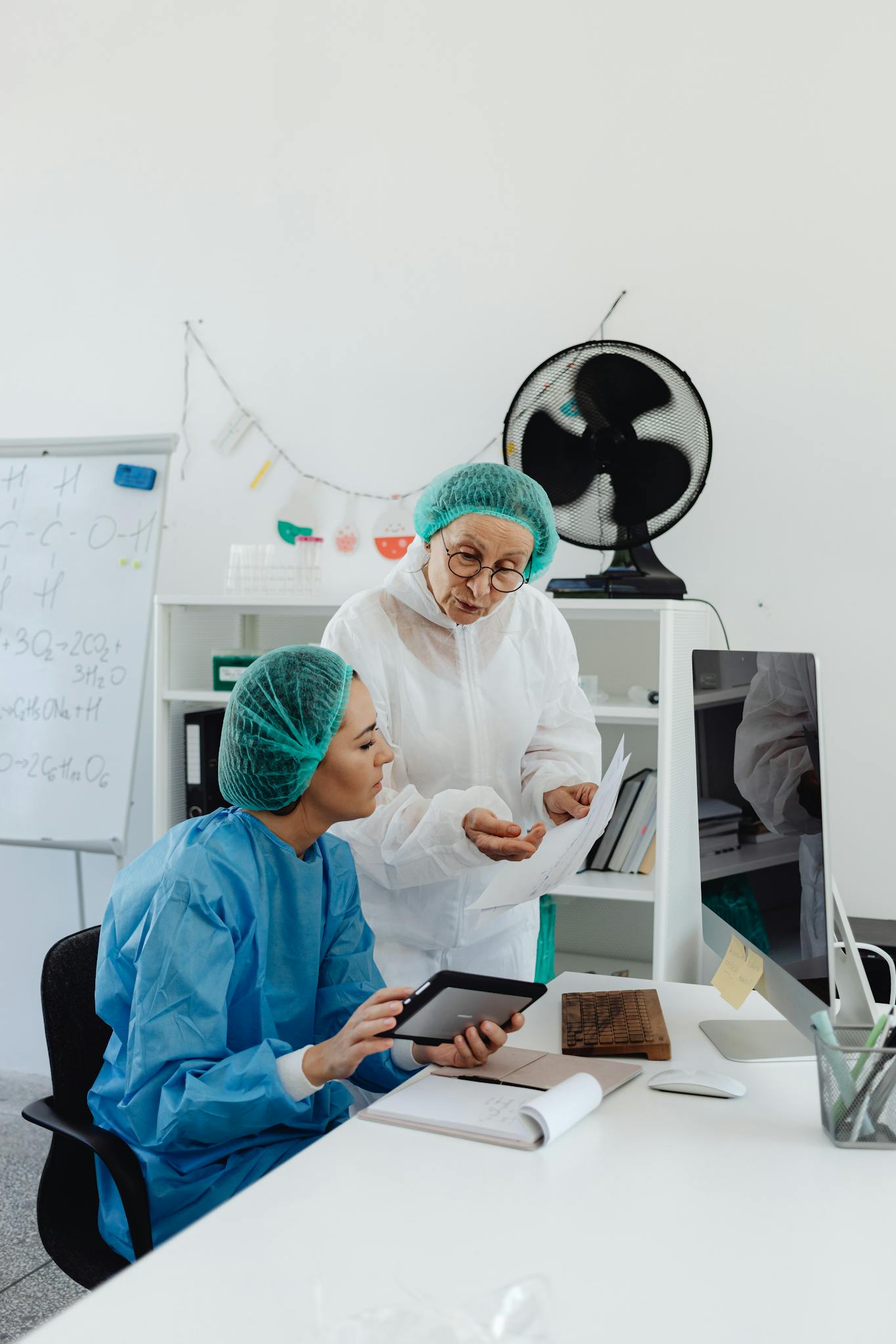 Two medical professionals in protective gear discuss research on a tablet in a laboratory setting.